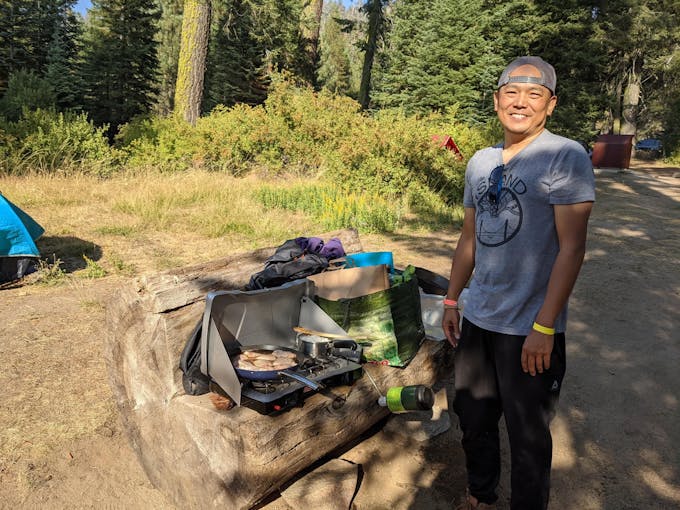 An Asian man wearing a trucker cap backwards and cooking chicken adobo on a campstove at a campsite with evergreen trees in the background.