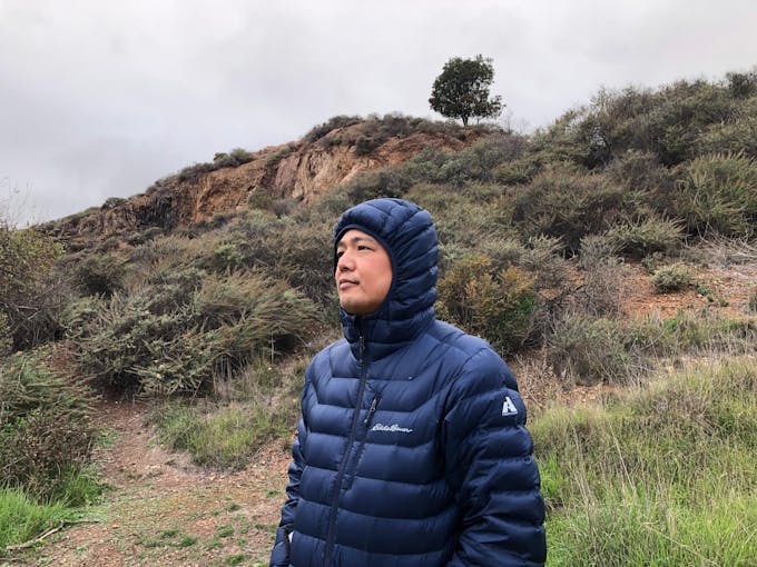A middle-aged Asian man wearing a blue hooded, puffy jacket looking out into the distance with plants and rocks in the background.