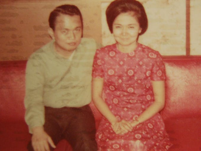 An Asian man and woman in 1960s-70s era clothing and hairstyles sitting close together on a bright red leather couch.