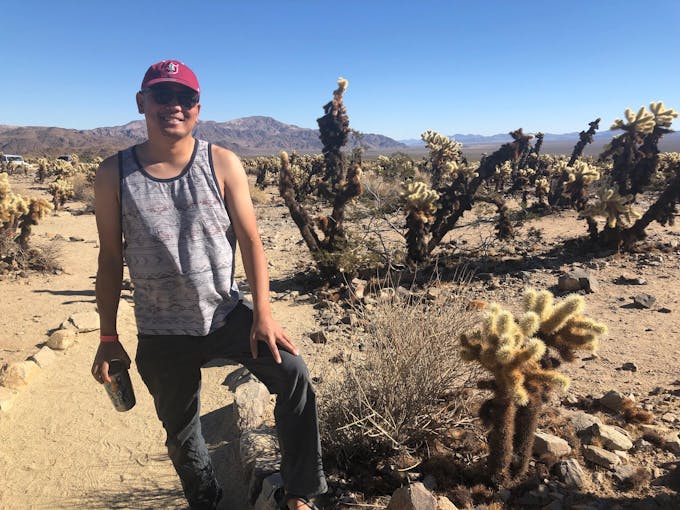 An Asian man wearing a tank top and a Stanford Cardinal cap with Cholla cacti behind them.