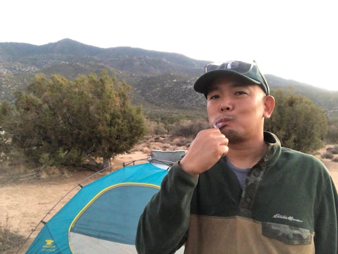 Asian man brushing their teeth with a bright blue tent behind them and green trees and mountains in the background.