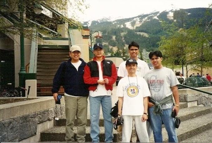 Five young Asian men standing on outdoor steps with melted ski runs in the mountains behind them.