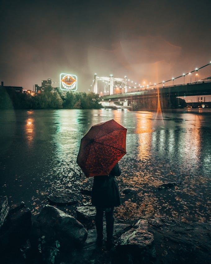 A person standing with a red umbrella on the Mississippi riverbank. It is nighttime and the giant neon Grain Belt Beer sign is lit up behind them.
