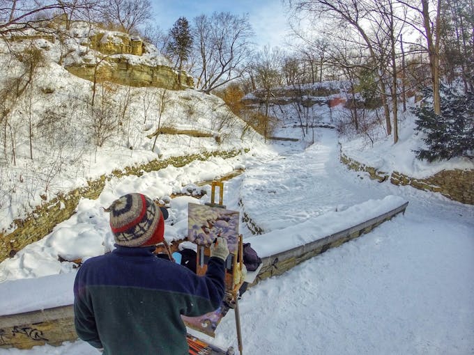 A person wearing a knit hat painting Minnehaha Falls on an easel on the walkway near the falls.