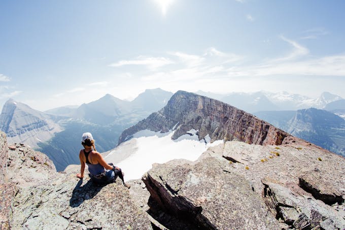 A woman sits at the edge of a mountain ridge with mountains in the background and bright sunlight.