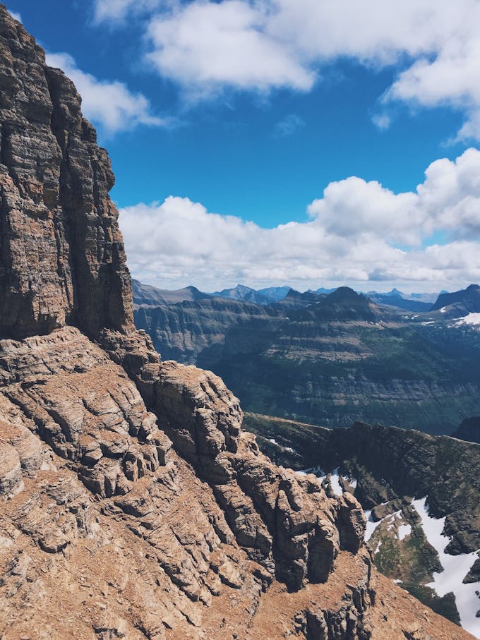 The side of a rocky mountain with other peaks in the distance. There is blue sky and clouds.