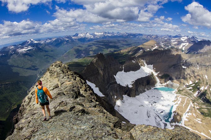 A person in a pink long-sleeve, shorts, and blue backpack walks along a rocky ridge over a snow-covered valley with blue sky and clouds.