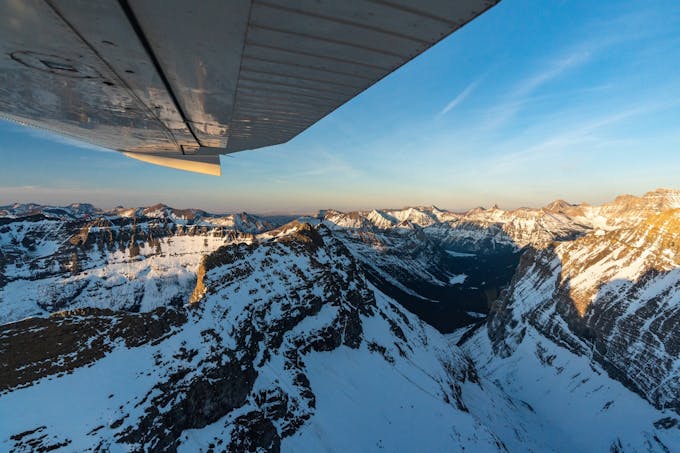 The view from a plane over Glacier National Park. The underside of the wing is visible and the mountains below are covered in snow.