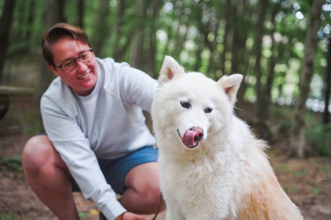 A person in a grey crewneck and blue shorts is kneeling down in the woods to pet a large, white, fluffy dog.
