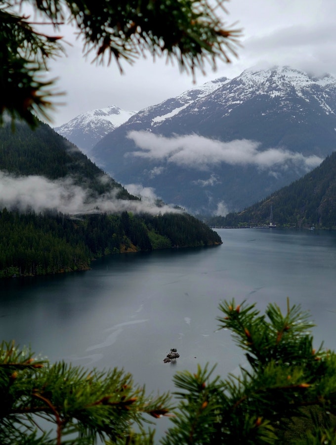 A through-the-trees view of an alpine lake surrounded by snow-capped mountains and whispy clouds.