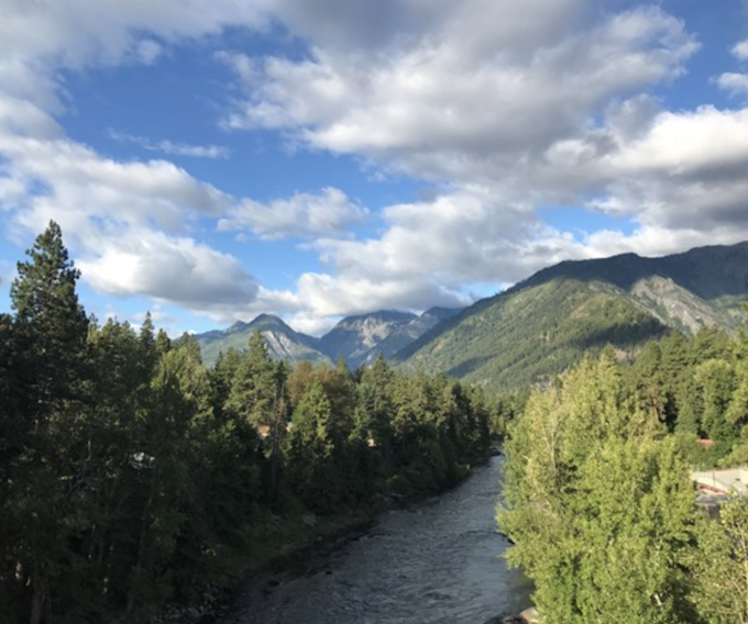 A river surrounded by pine trees with mountains in the background. The sky is baby blue with puffy clouds.