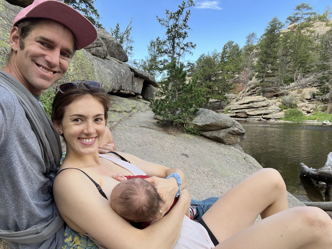 A young caucasian couple is sitting next to  a river. The man is holding the camera as he sits behind the woman. The woman is breastfeeding an infant. There are evergreen trees in the background.