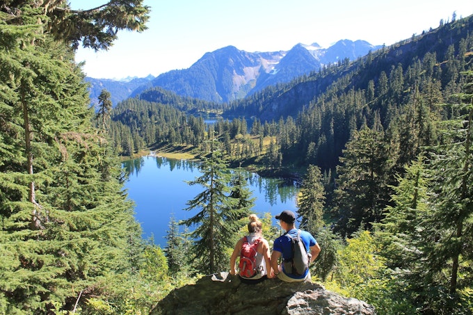 Two people sit on a rock face over an alpine lake. There are evergreen trees surrounding the lake and mountains in the background.