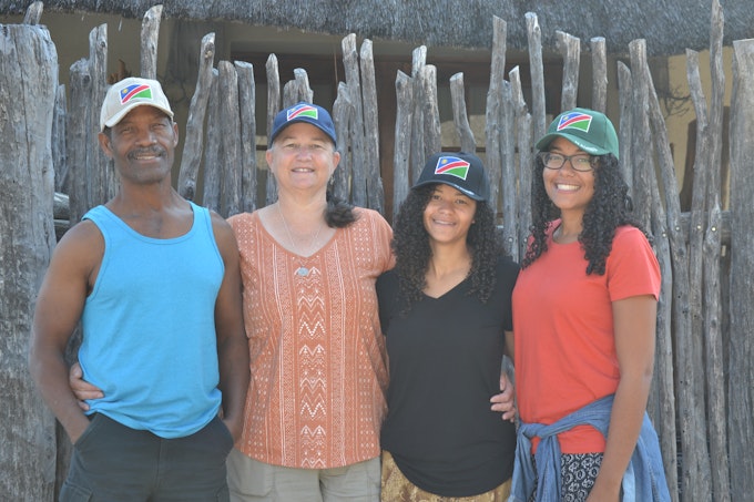 Father, mother and two daughters stand in front of wooden fence with hats that have Namibian flag on them.
