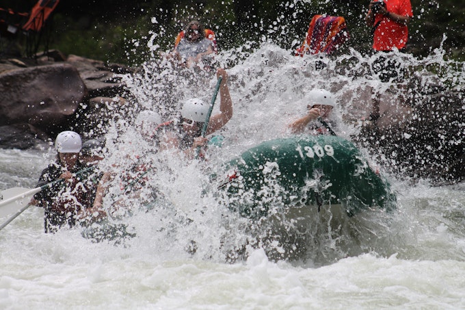 Three people in large green raft with white water splashing up around them.