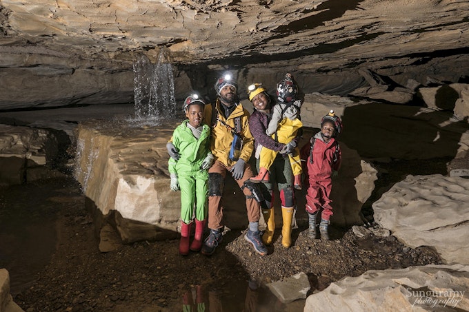 A black family of two adults and three young kids are seated in a cave. There is a waterfall behind them and they are wearing brightly colored clothes, helmets, and headlamps.