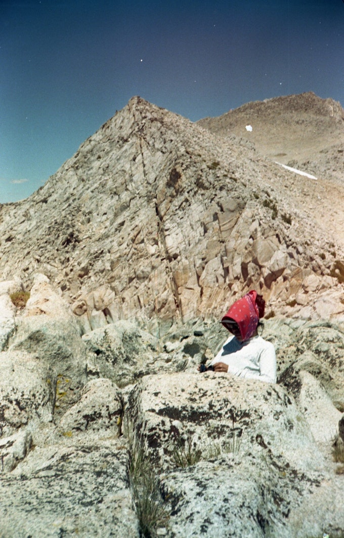 A person wearing a red bandana and a white, long-sleeved shirt sits behind a granite rock and very closely in front of a High Sierra peak. The sky is a deep, clear blue.