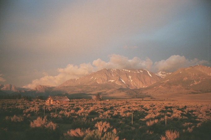Clouds sit just above Sierra Nevada mountains, colored red by the rising sun. There is a wooden, old building sitting wtihin the sagescrub desert floor.
