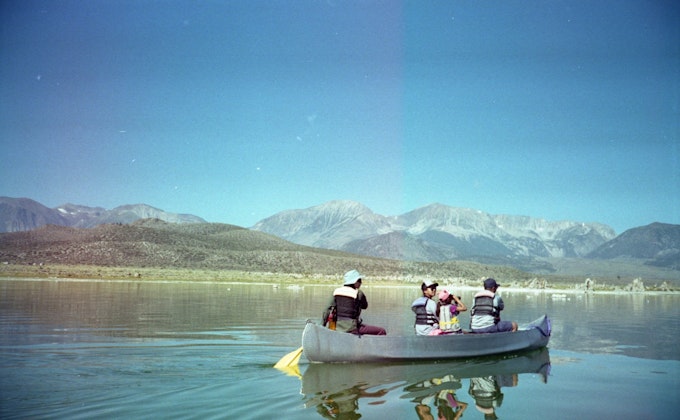 Four people sit in a gray canoe floating on still water. The sky is blue and clear above the Sierra Nevada mountain range.