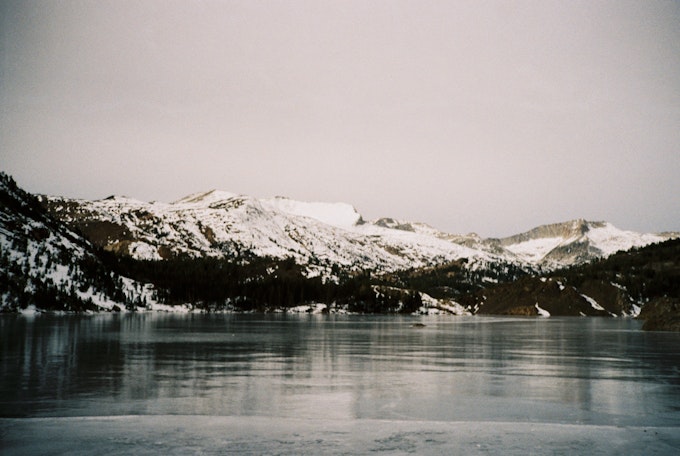 Snowy mountains sit just behind an unfrozen lake. Gray skies sit above.