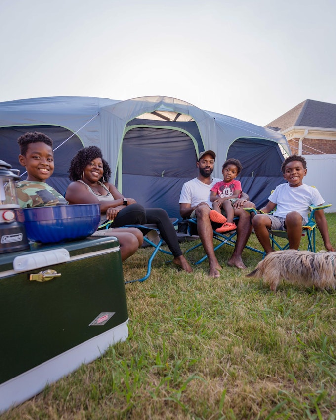 Two parents and three little kids sitting on lawn chairs in front of a large tent. There is a small shaggy dog in the grass.
