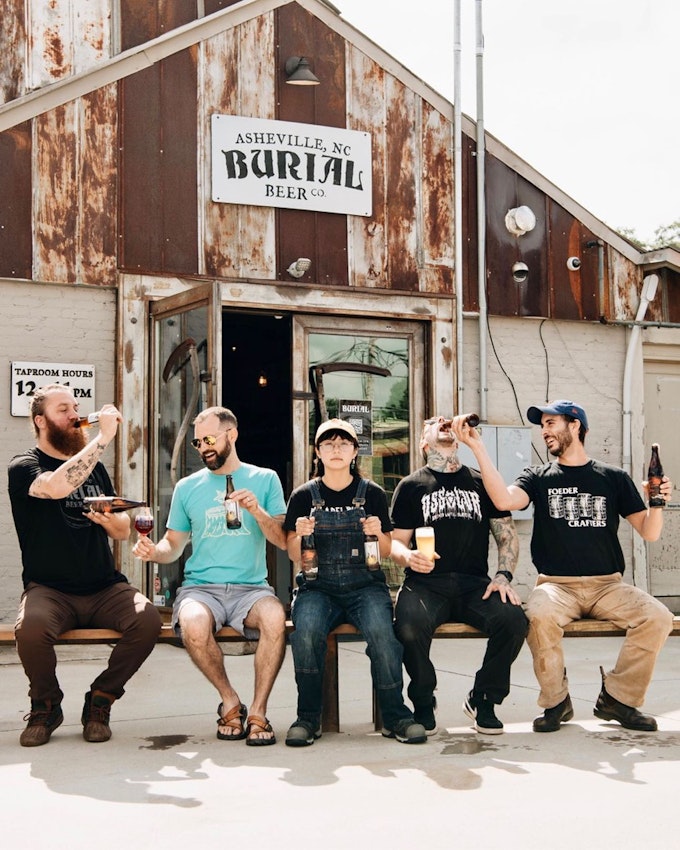 Five people are seated on a bench in front of a building. They are drinking beer and enjoying the sunshine.