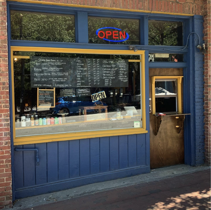 The exterior of a coffee shop. It has blue wood paneling and yellow window trim around a window that shows a menu and some drink options inside. A neon open sign is lit up above the window and the door has a window that opens to provide service to customers outside.