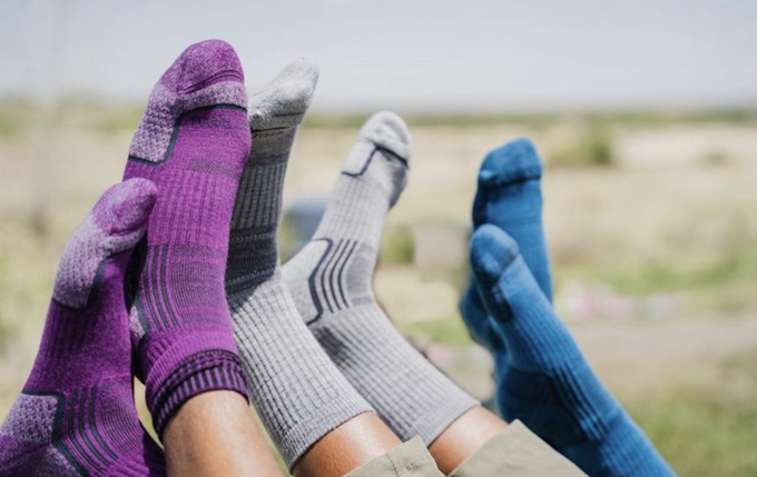 Three people's feet are up in the air. One is wearing purple socks, another grey, and another blue. There is grass in the background.