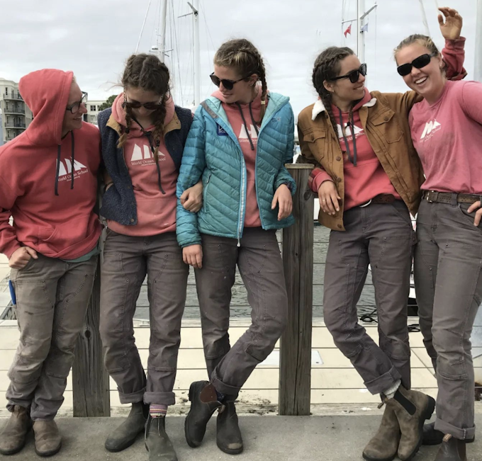 A group of people with side braids wearing matching grey work pants and Blundstone boots. They wear pink sweatshirts and have their arms around each other on a dock or wharf near water.