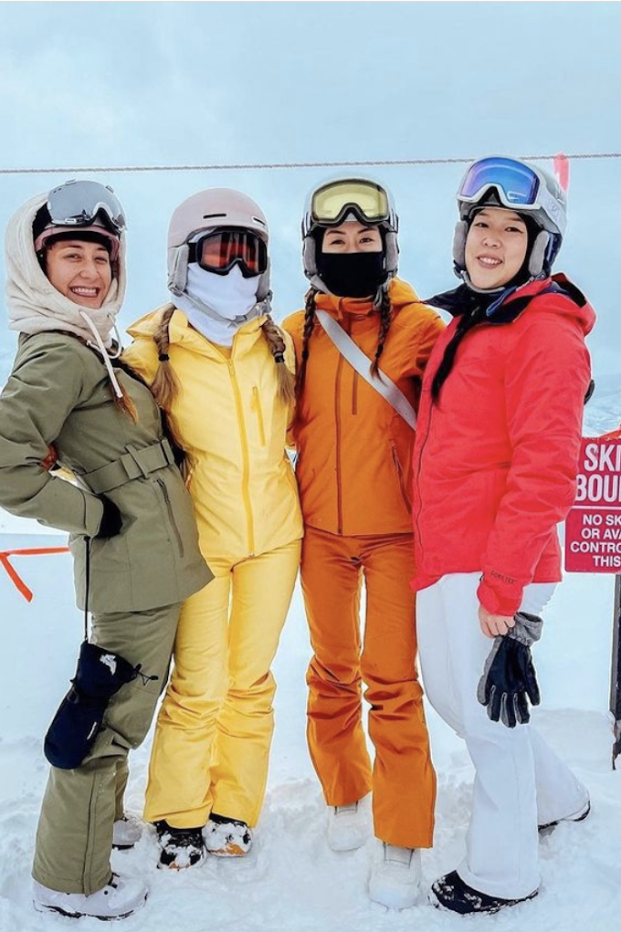 Four women stand in colorful ski outfits. They are smiling at the camera and wearing helmets and goggles. They are standing on snow next to a sign that reads, 