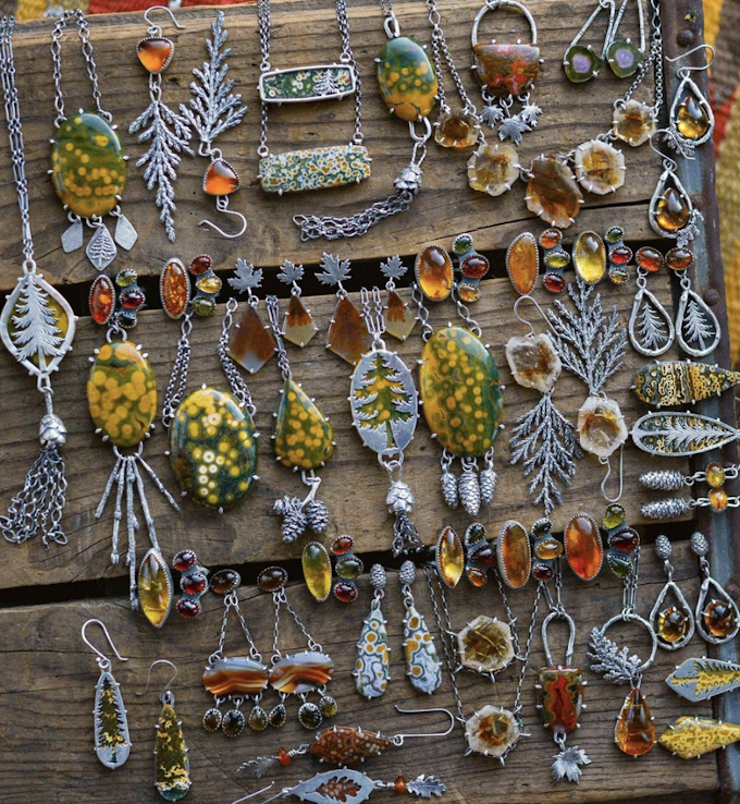 A from-above shot of a lot of silver jewelry laid out on rustic wood. The jewelry includes earrings and necklaces made from silver and yellow, green, orange, and red gems and stones. There are silver cutouts of pine needle leaves, trees, pinecones, and leaves.