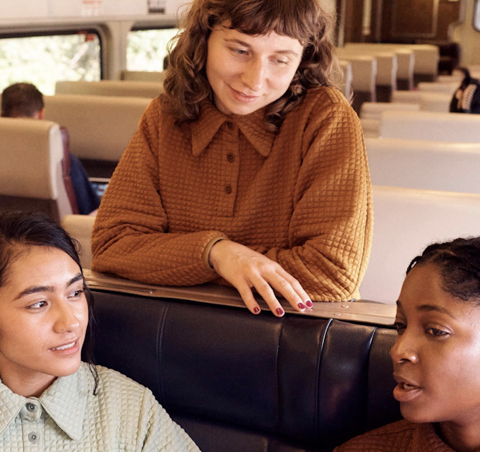 three people of different ethnicities are seated on a bus. Two are sitting together and the third is leaning over the seat ahead to talk.