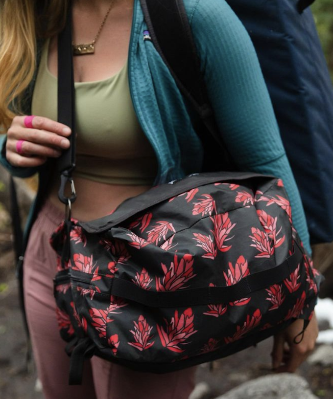 A person wearing pink pants, a green sports bra and teal sweatshirt is carrying a black and pink floral sports bag.