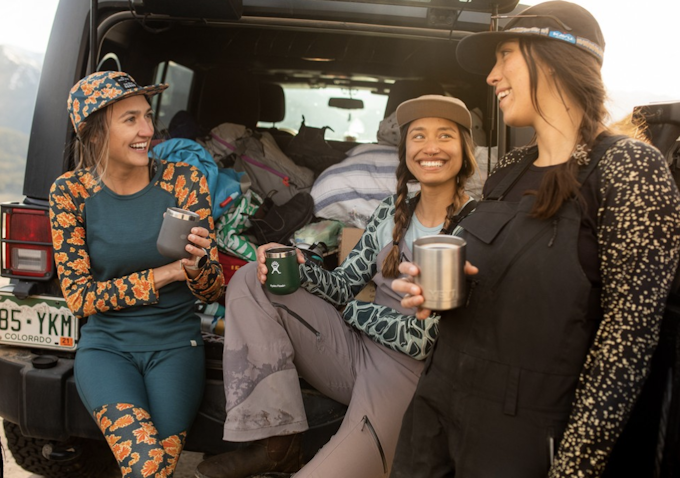 Three people are wearing colorful patterned clothing and sipping beverages from thermoses. They are seated on the bed of a truck as if they just got done with an outdoor activity.