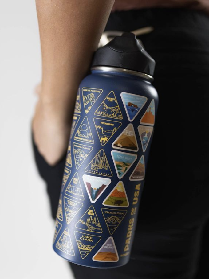 A close-up image of a blue water bottle attached to someones pants belt loop. Their hand is in their pocket behind the bottle and out of focus. The bottle has empty triangles representing national parks the owner has not visited. Some are filled in with stickers from the parks they have visited.