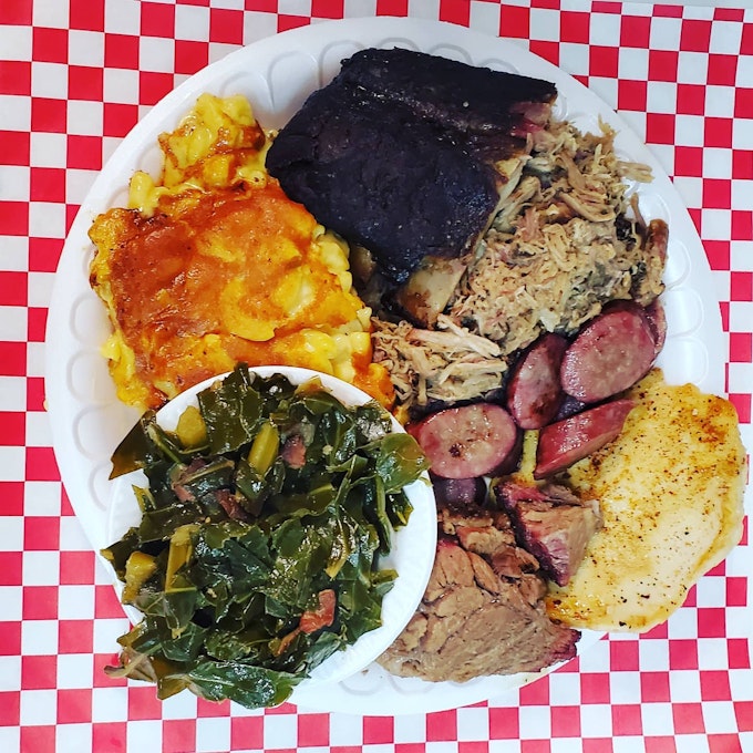 A from-above view of a white plate set on a checkered table cloth. The plate hosts various meats, greens, and eggs.