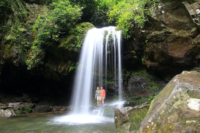 Two people stand behind a wispy waterfall topped with greenery and surrounded by rocks.