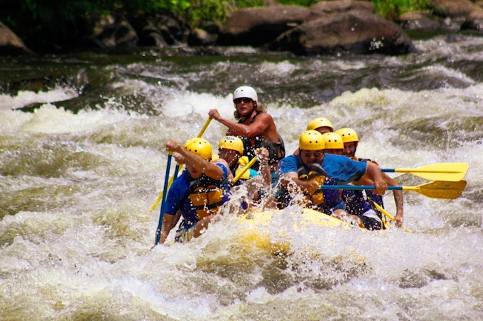 A group of paddlers in yellow helmets paddle a yellow raft through a huge whitewater set.