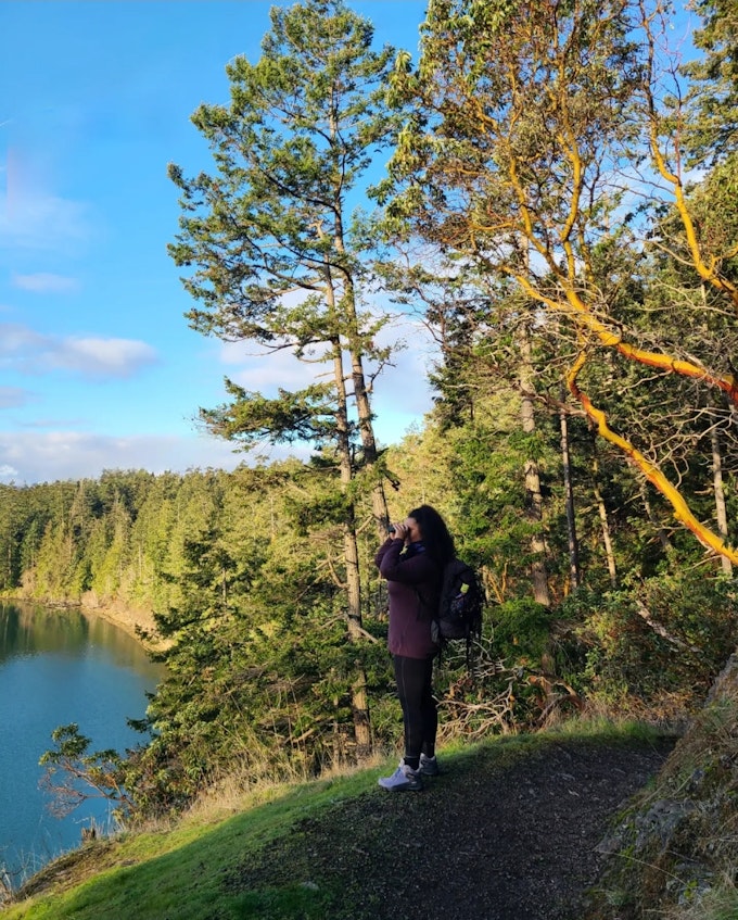 A person is standing on a mossy cliff looking out over water with binoculars.