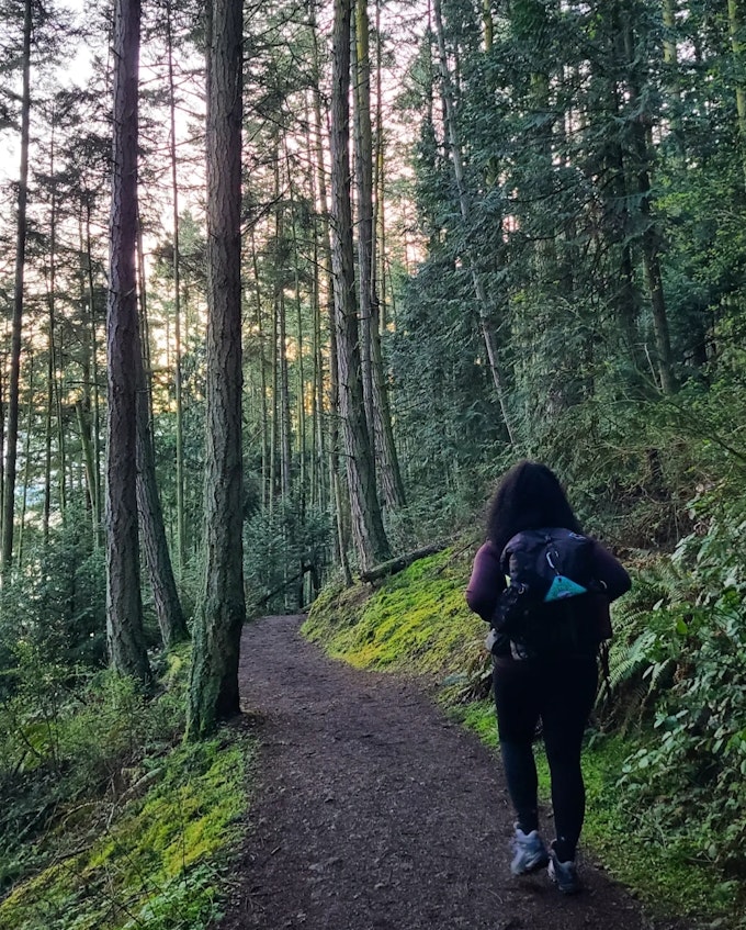 A person with dark black hair, a purple shirt, black leggings, and hiking boots is wearing a hiking pack and walking away from the camera in a lush forest.