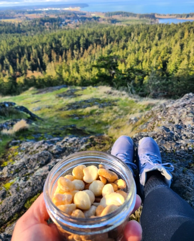 A person is holding a jar of nuts while seated on a cliff over a forest. There is a body of water in the distance.
