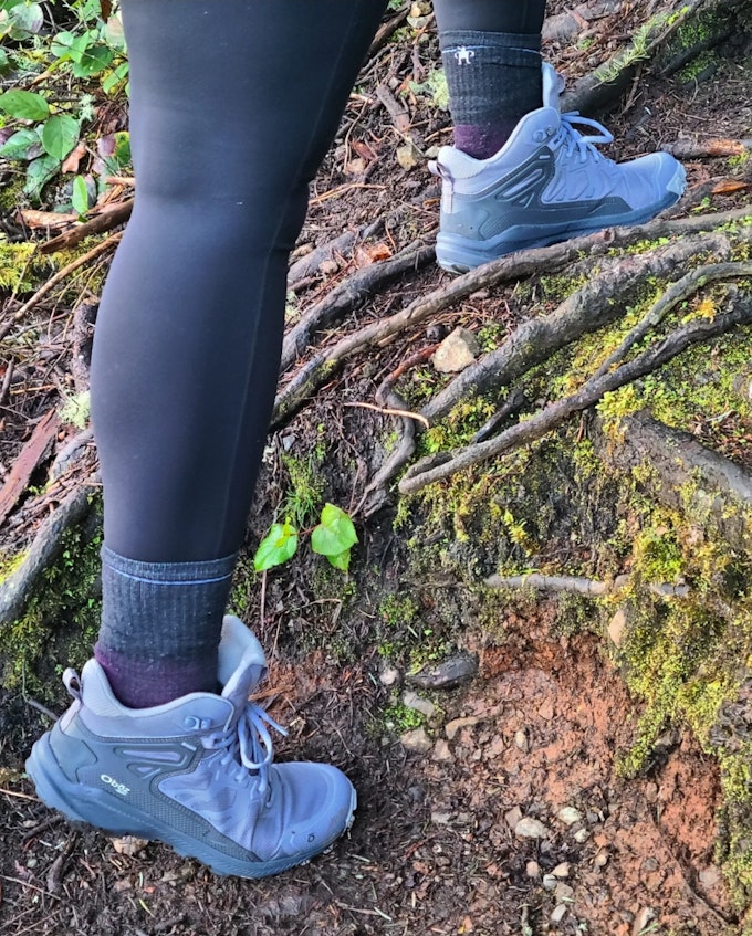 A close-up of a hiker wearing leggings and purple hiking boots while walking over rooty moss.