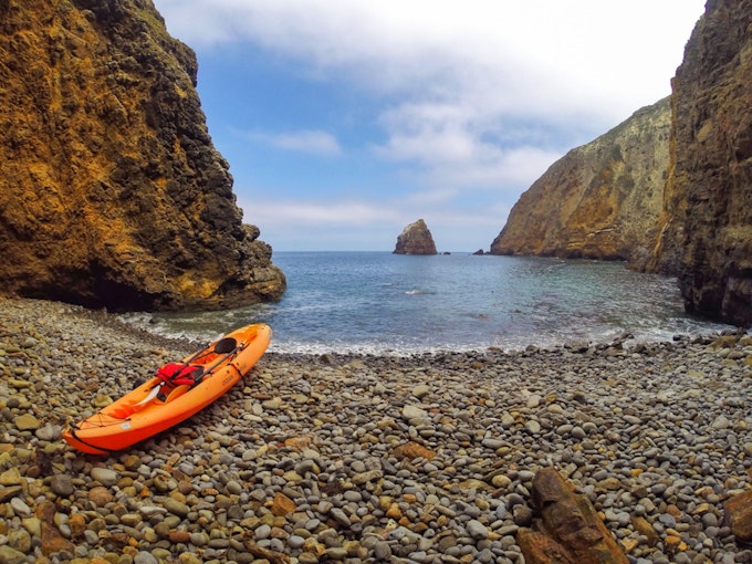 Orange kayak sitting on the rocky shore of a blue ocean. A rock formation sits out in the distance under a cloudy blue sky in Channel Islands National Park.
