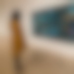 A black person in a white jacket, leggings, and shoes is standing on light wood floor looking at a large painting on a white wall. The painting features people in blues, blacks, and yellows.