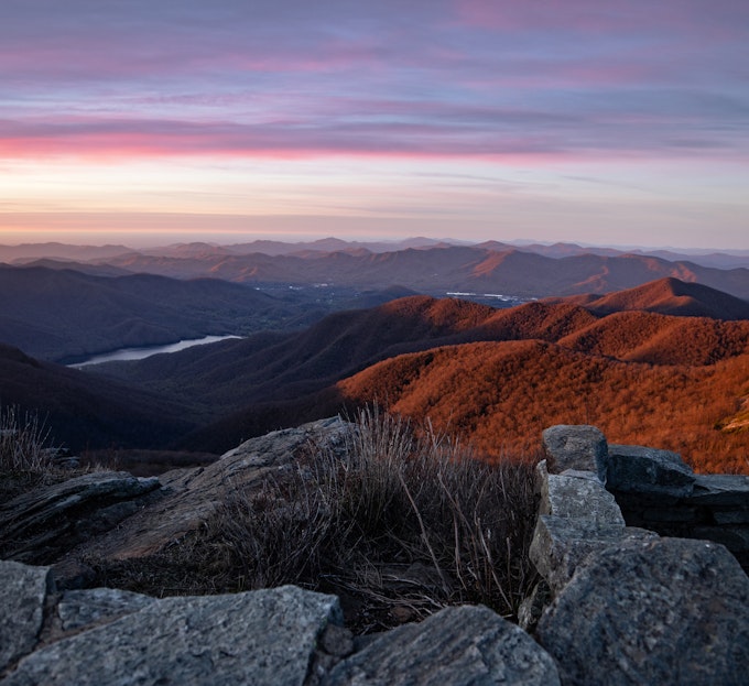 The sun is rising over mountains and a river. The mountains have an orange glow and the sky is light blue, pink, and purple.