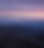 Rounded mountains reach from one side of the image to the other. The closer ones are dark and the further ones are a lightening purple. The sky is purple, blue, pink, orange, and yellow as the sun rises.