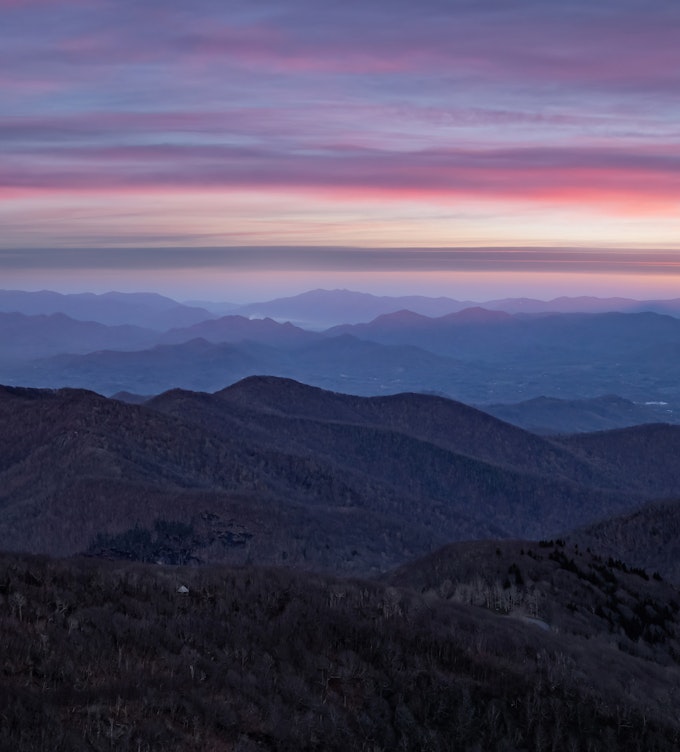 Rounded mountains reach from one side of the image to the other. The closer ones are dark and the further ones are a lightening purple. The sky is purple, blue, pink, orange, and yellow as the sun rises.
