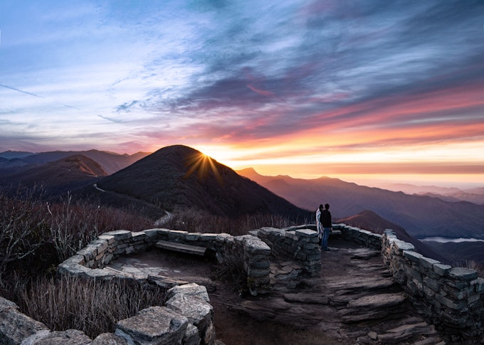 A rock wall winds to an overlook where two people stare out at a sunrise. The sky is many colors and the sun is barely up high enough to see over a mountain peak.