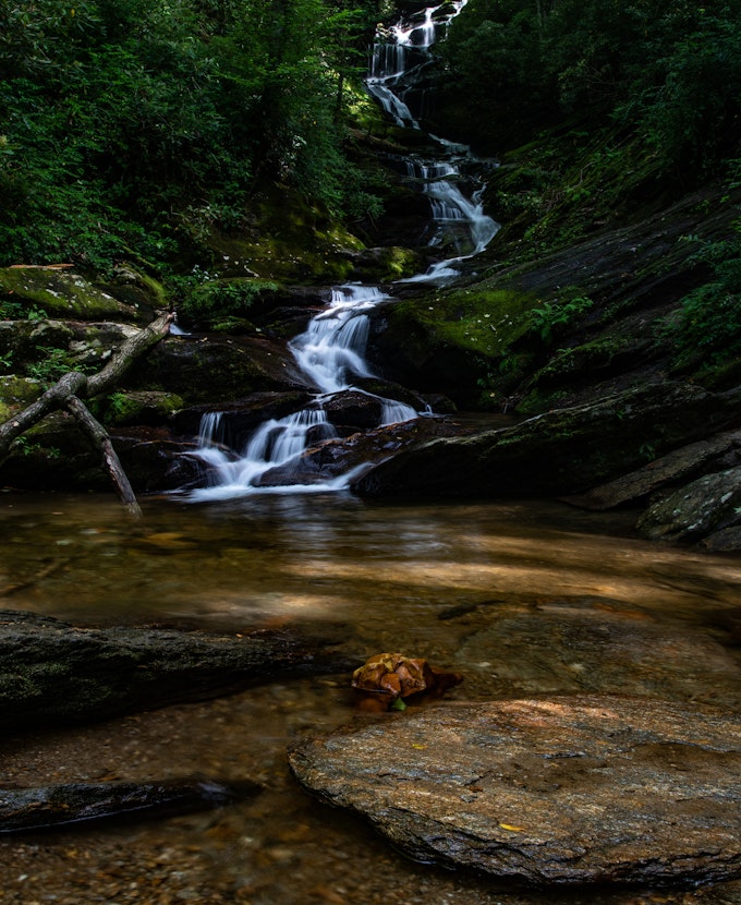 A tall, thin waterfall trickles over piles of rocks and greenery and into a clear pool.
