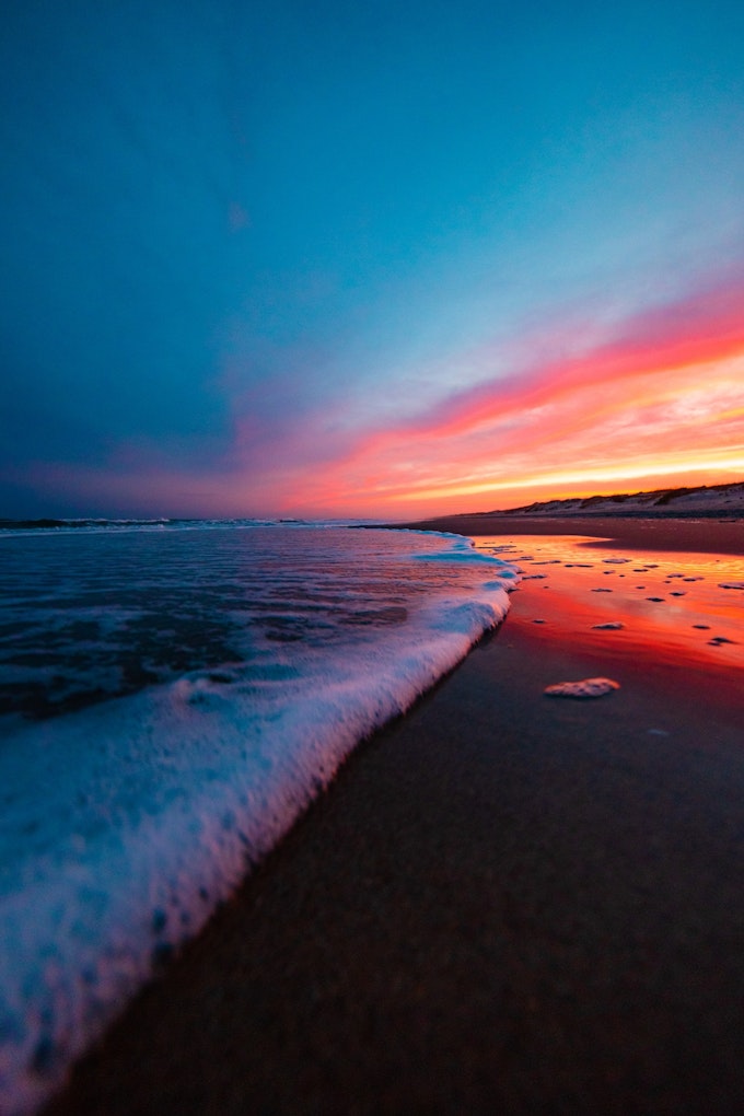 The camera is near ground-level on a beach as a frothy wave rolls in. The sun is setting in the distance and the sky is a dark blue fading into purple, pink, orange, and yellow.